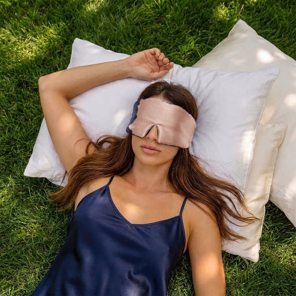 Frau mit der Luxus Lyocell Schlafmaske in Light Rose auf wei�en Kissen im Gras